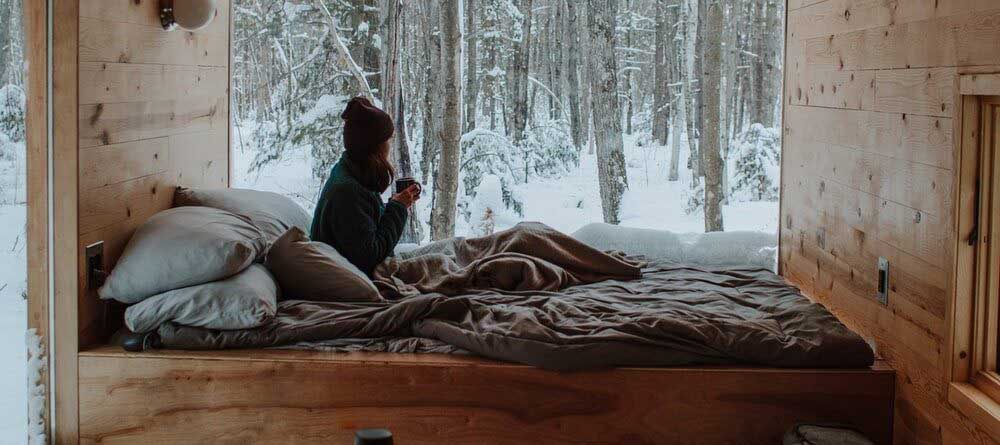 A person sitting on bed with a cup of coffee looking into frozen backyard